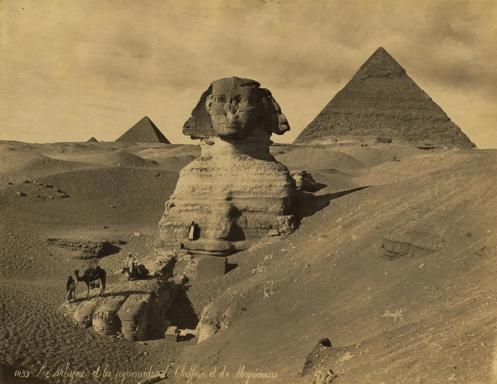 An old sepia photograph of the Sphinx half buried in sand, with two pyramids behind it.